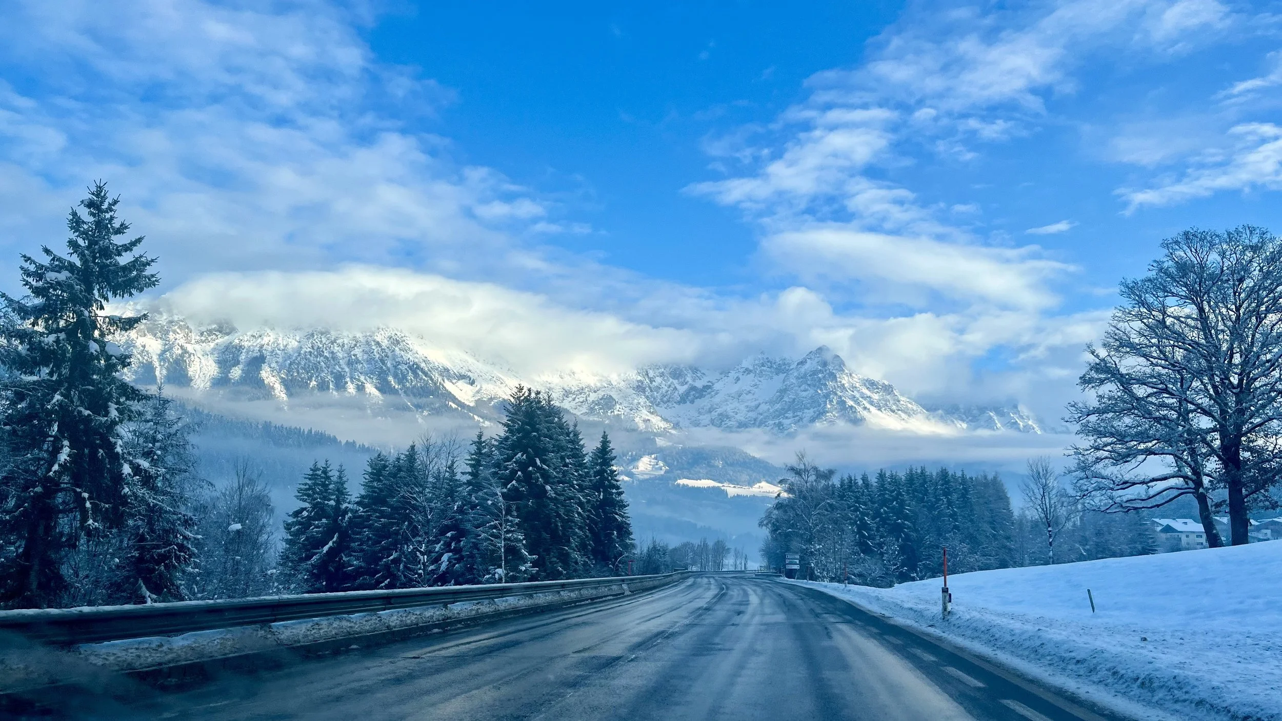Eine schneebedeckte Straße führt durch eine winterliche Landschaft mit Nadelbäumen, Bergen im Hintergrund und einem blauen Himmel mit Wolken.