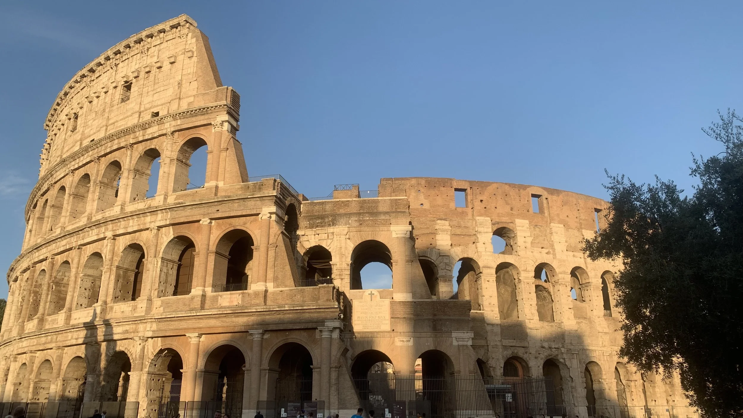 Das Kolosseum in Rom bei Sonnenuntergang, sichtbar mit mehreren Bögen und einer Ecke, umgeben von Menschen und Bäumen.