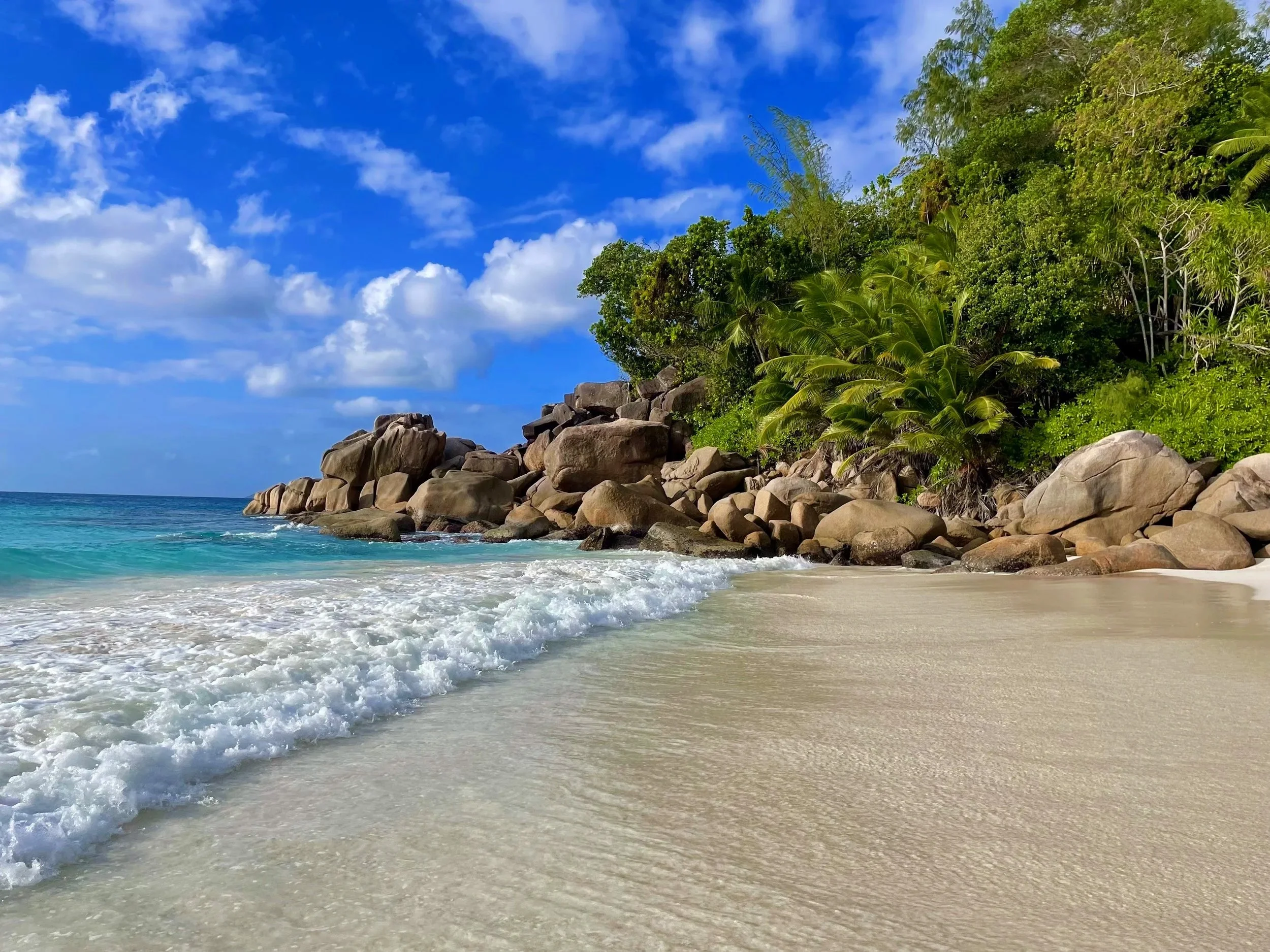 Strand mit weißen Sand, Wellen und grünem Dschungel mit Felsen im Hintergrund unter blauem Himmel mit Wolken.