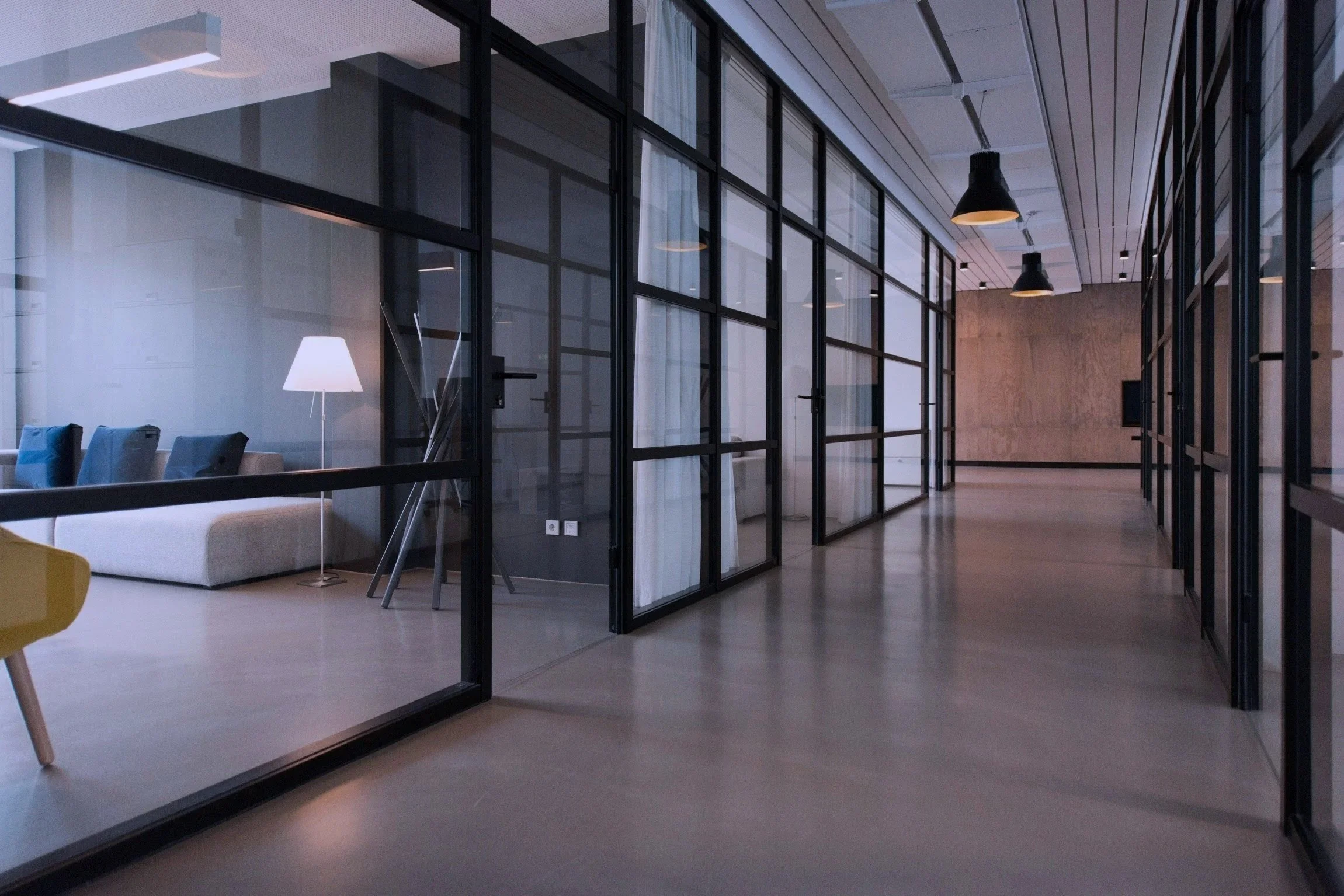 Modern office with glass walls, wooden flooring, black pendant lighting, and a sitting area with a sofa and a lamp.