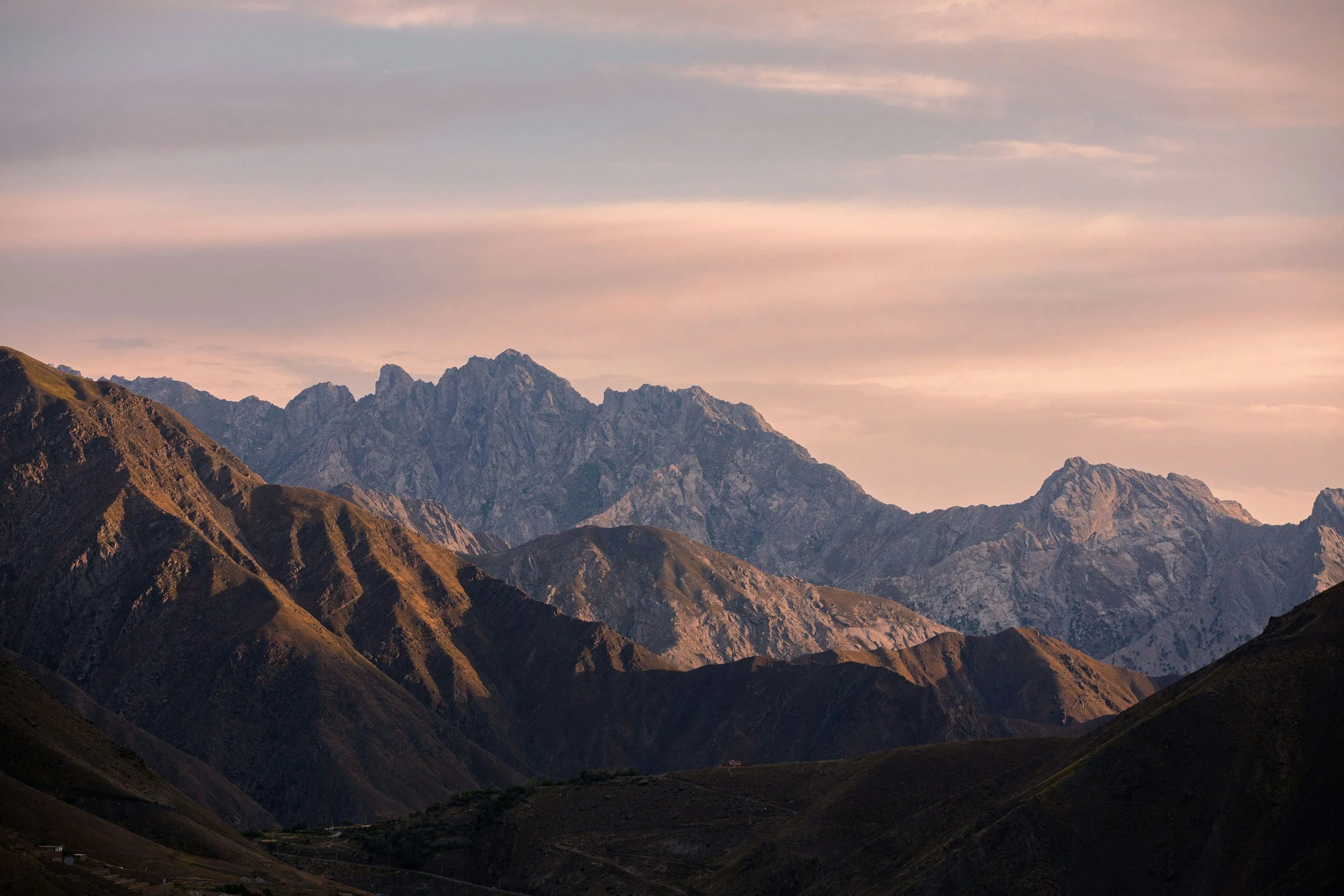 Mountain range with rugged, rocky peaks and rolling hills under a cloudy sky during sunset.