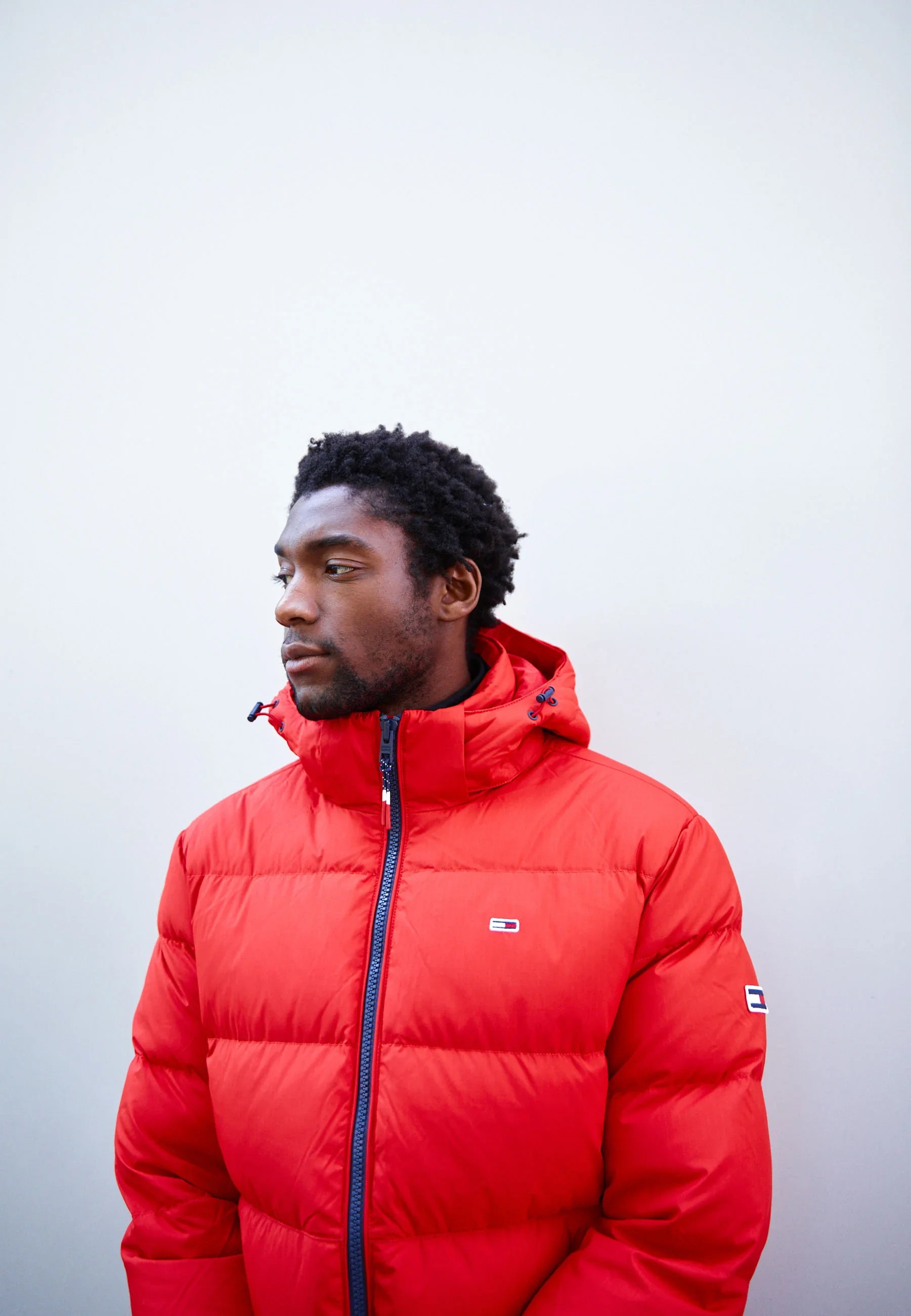 A man in a red puffer jacket with a hood standing against a plain white wall, looking slightly to the side.
