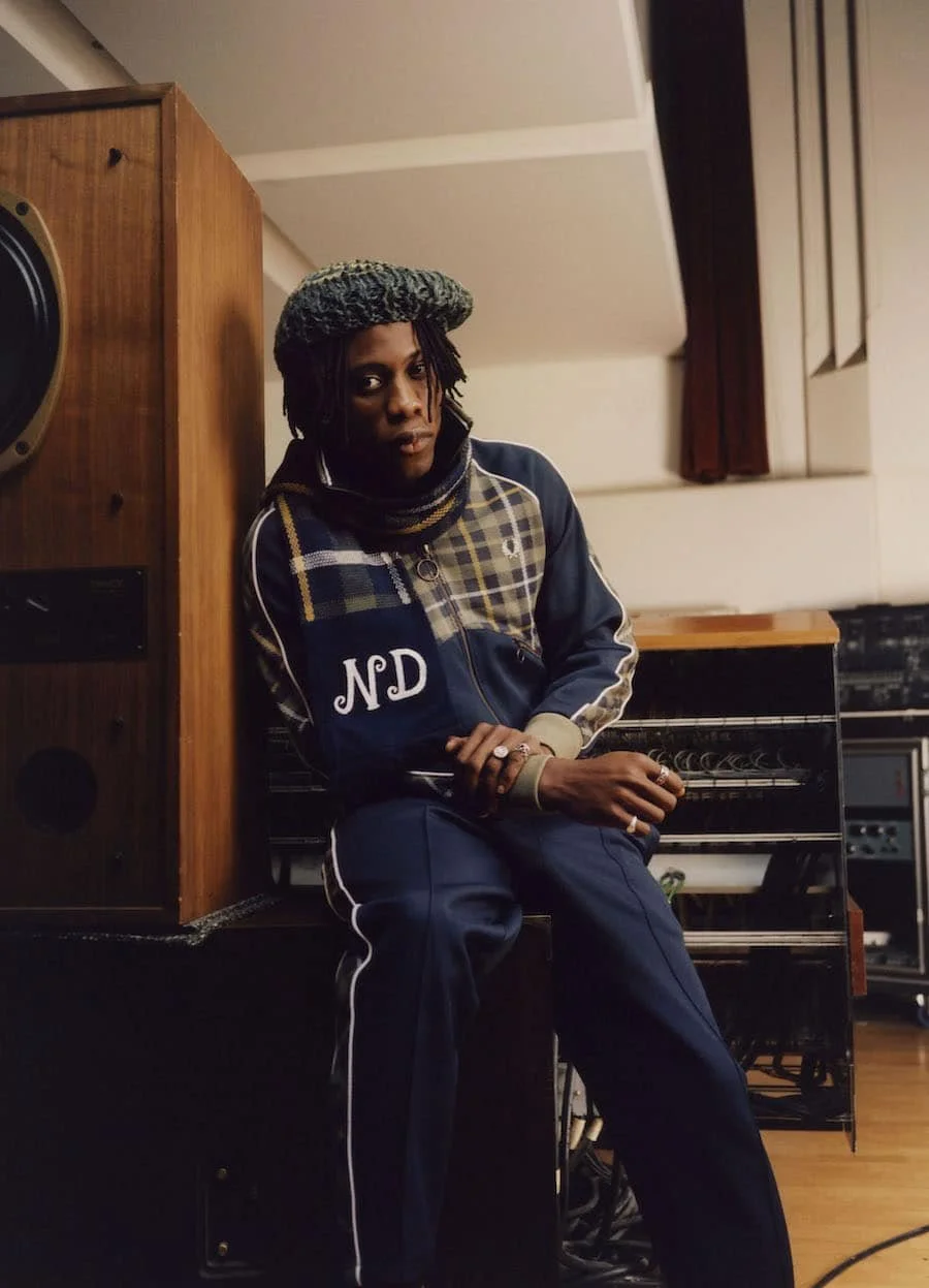 A person with dark skin, wearing a patterned beret and a sports jacket sitting on a bench in a room with musical equipment, including a piano and a large speaker.
