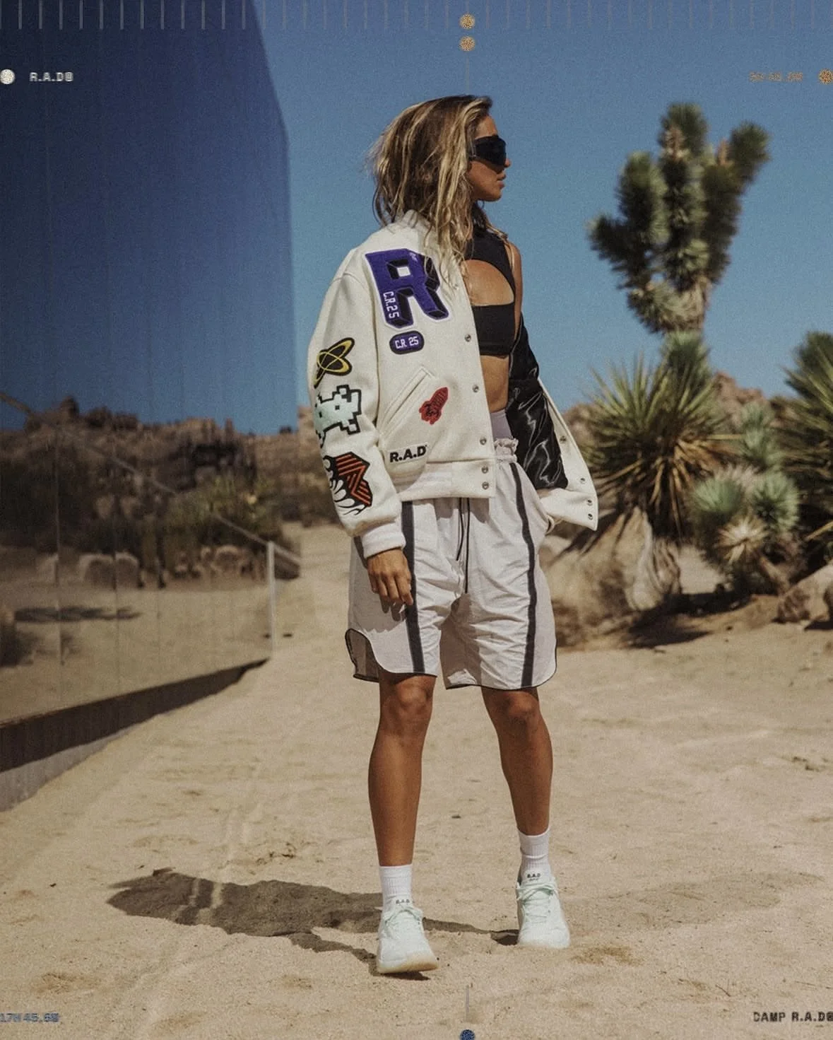 A woman standing outdoors in a desert landscape, wearing a black crop top, white shorts with black stripes, white sneakers, and a white jacket with colorful patches. She has long, wavy blonde hair and wears a black eye mask, with desert plants and a clear blue sky in the background.