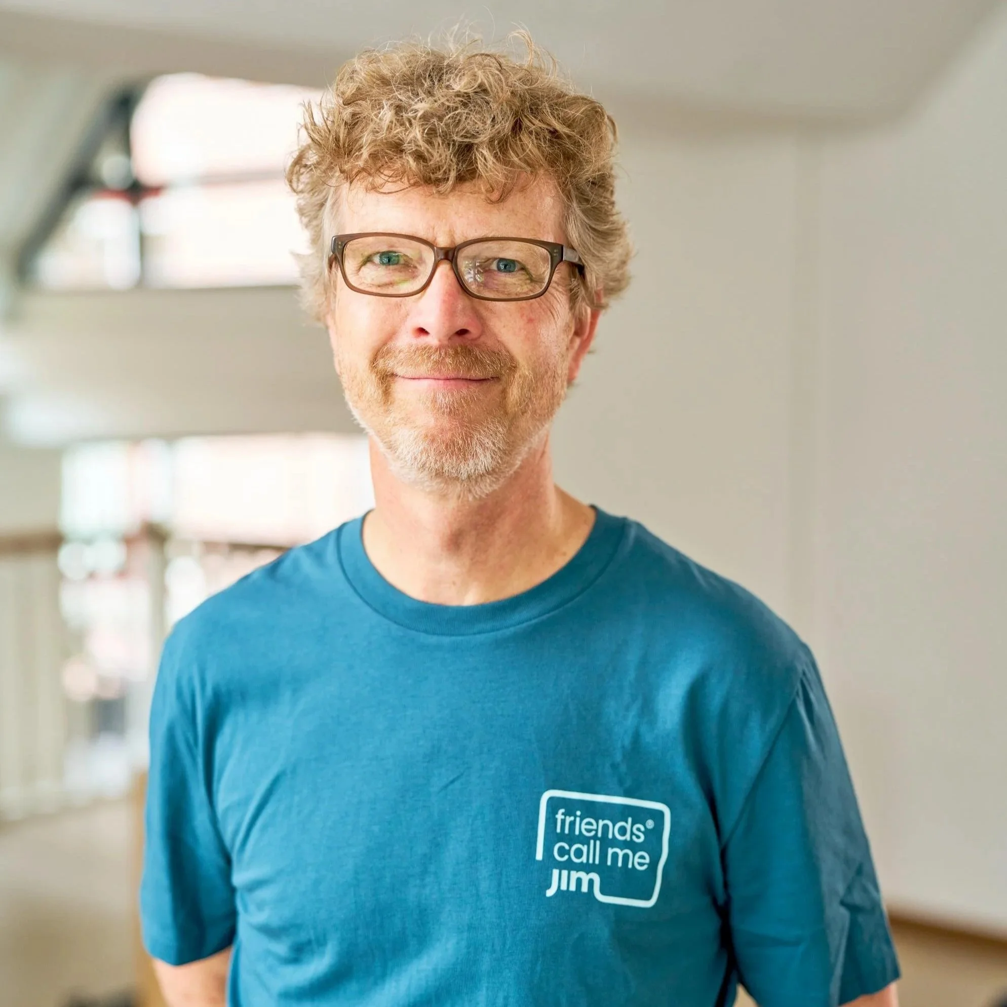 Man met bril, geschild haar en een baard, draagt een blauwe T-shirt met tekst 'friends call me Jim', glimlacht in een lichte ruimte.