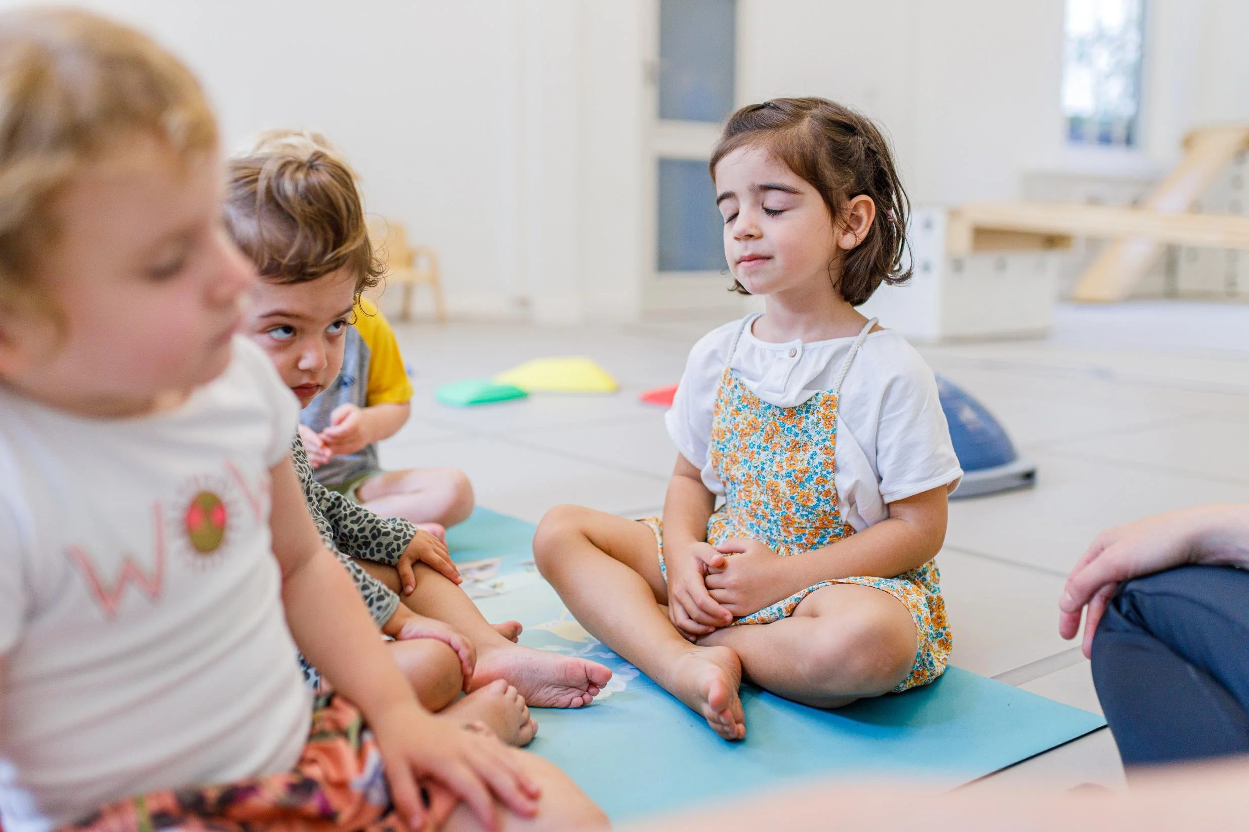 Meisje zit op een yogamat met haar ogen dicht, omringd door andere kinderen in een lichte speellocatie.