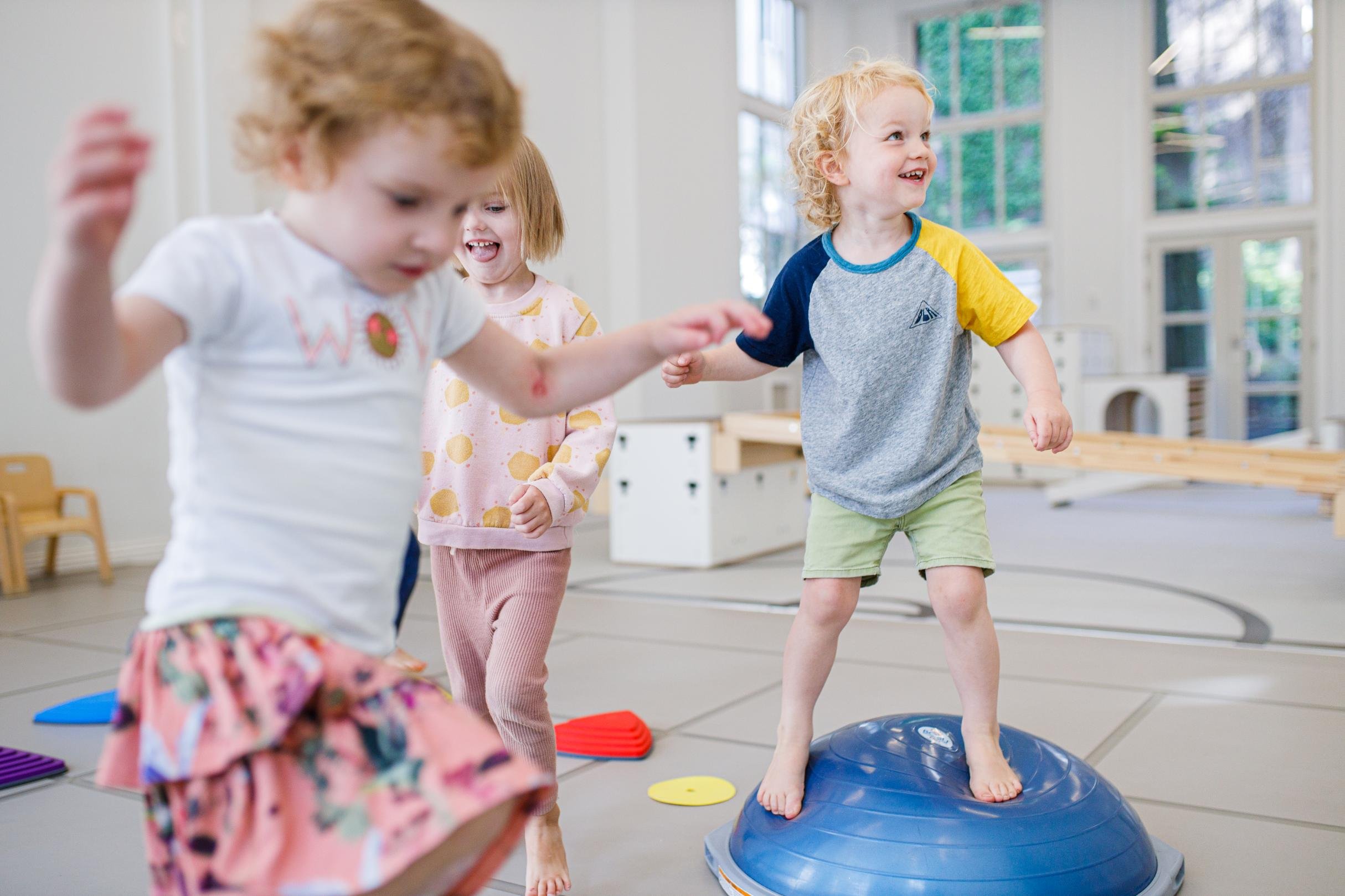 Drie kinderen die plezier hebben in een binnenruimte, met één kind die op een blauwe oefenbal staat en twee anderen die eromheen spelen.
