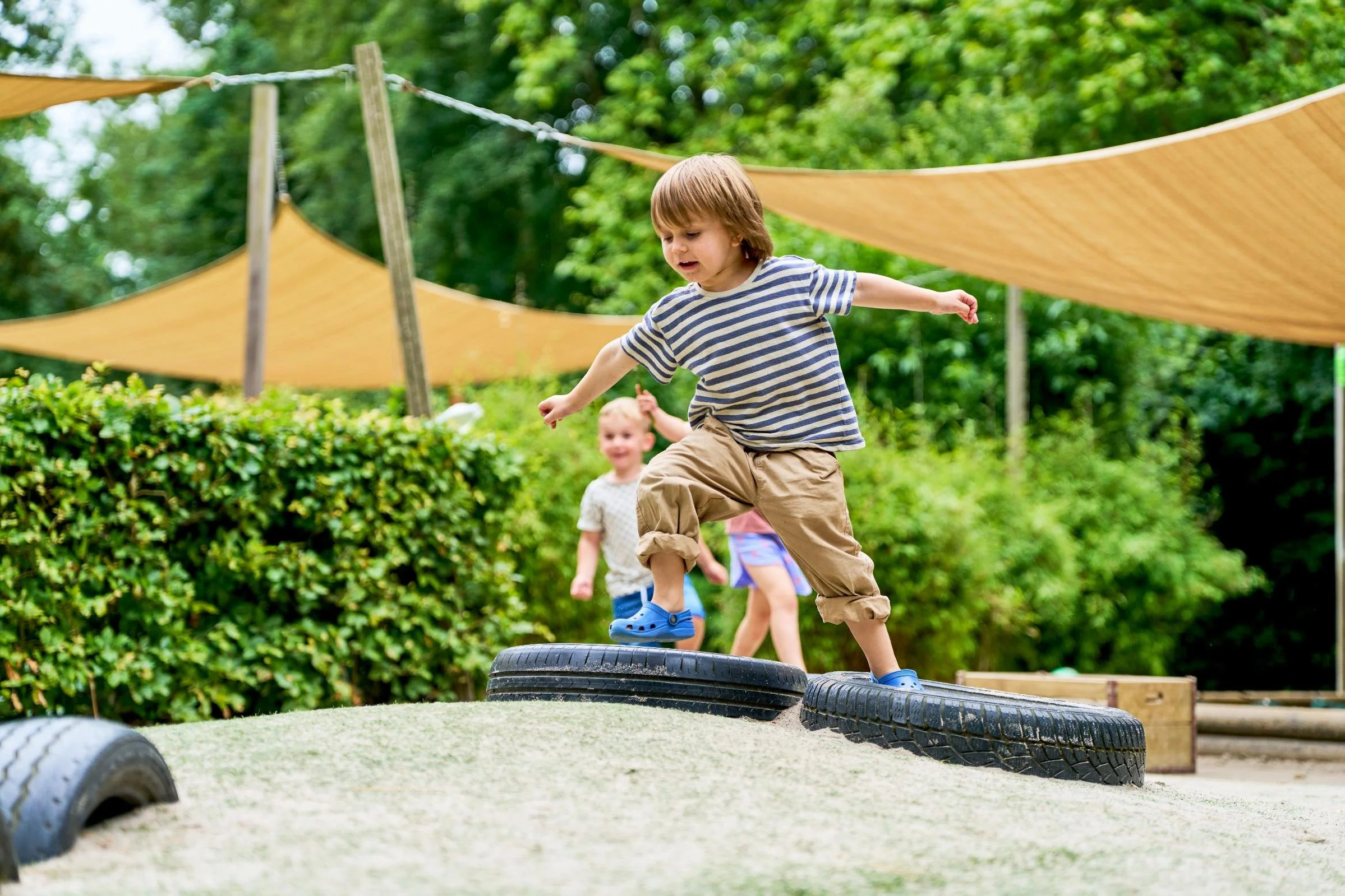 Kinderen die op een buitenspeeltuin met oude banden balanceren, omgeven door groene bomen en zonwerende doeken.
