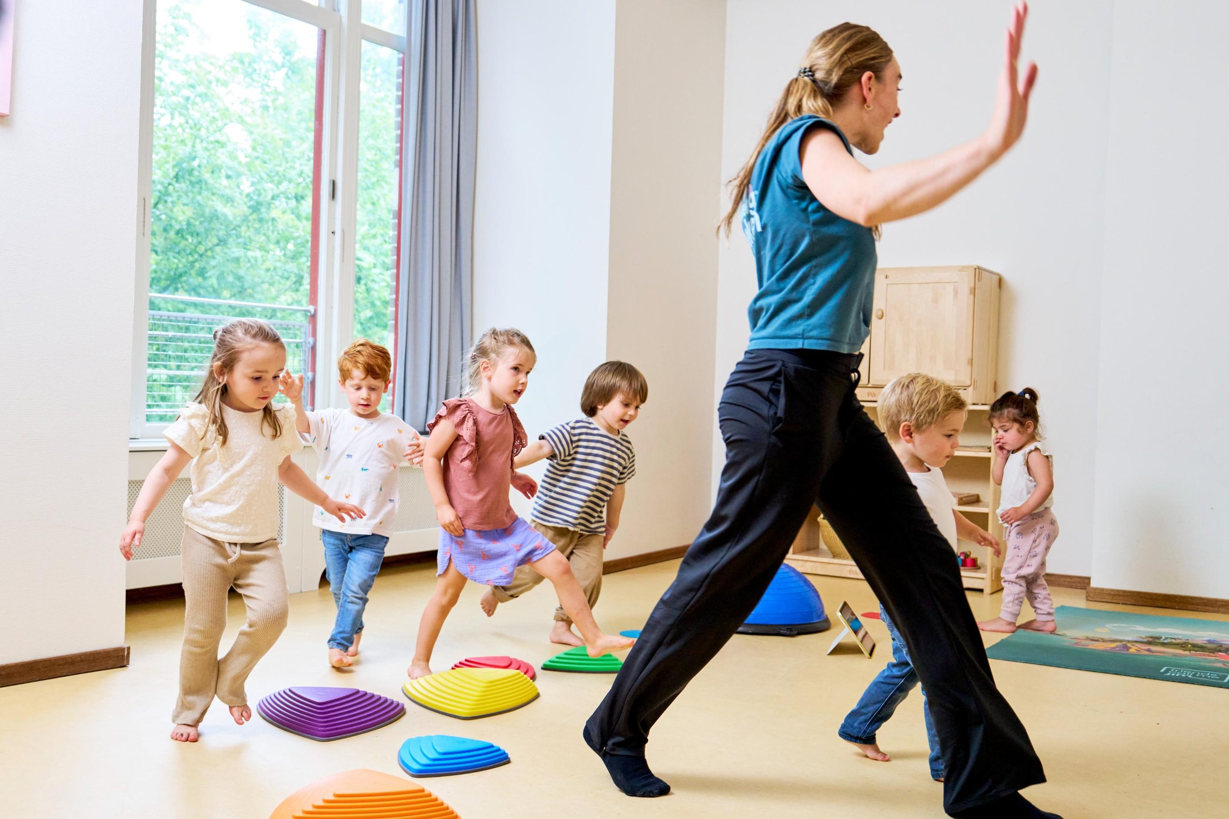 Een vrouw leidt een groep jonge kinderen in een indoors oefening of spel, waarbij ze over kleurrijke opstapjes lopen.