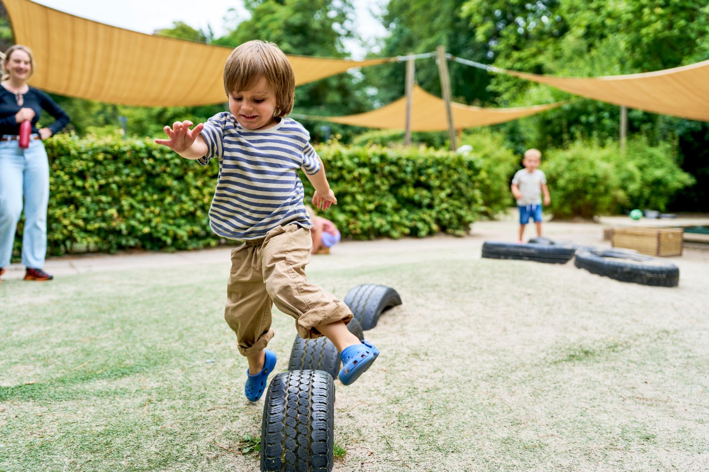 Kind speelt op een bandenslipbaan in een speeltuin, omringd door struiken en bomen, met volwassenen en andere kinderen op de achtergrond.