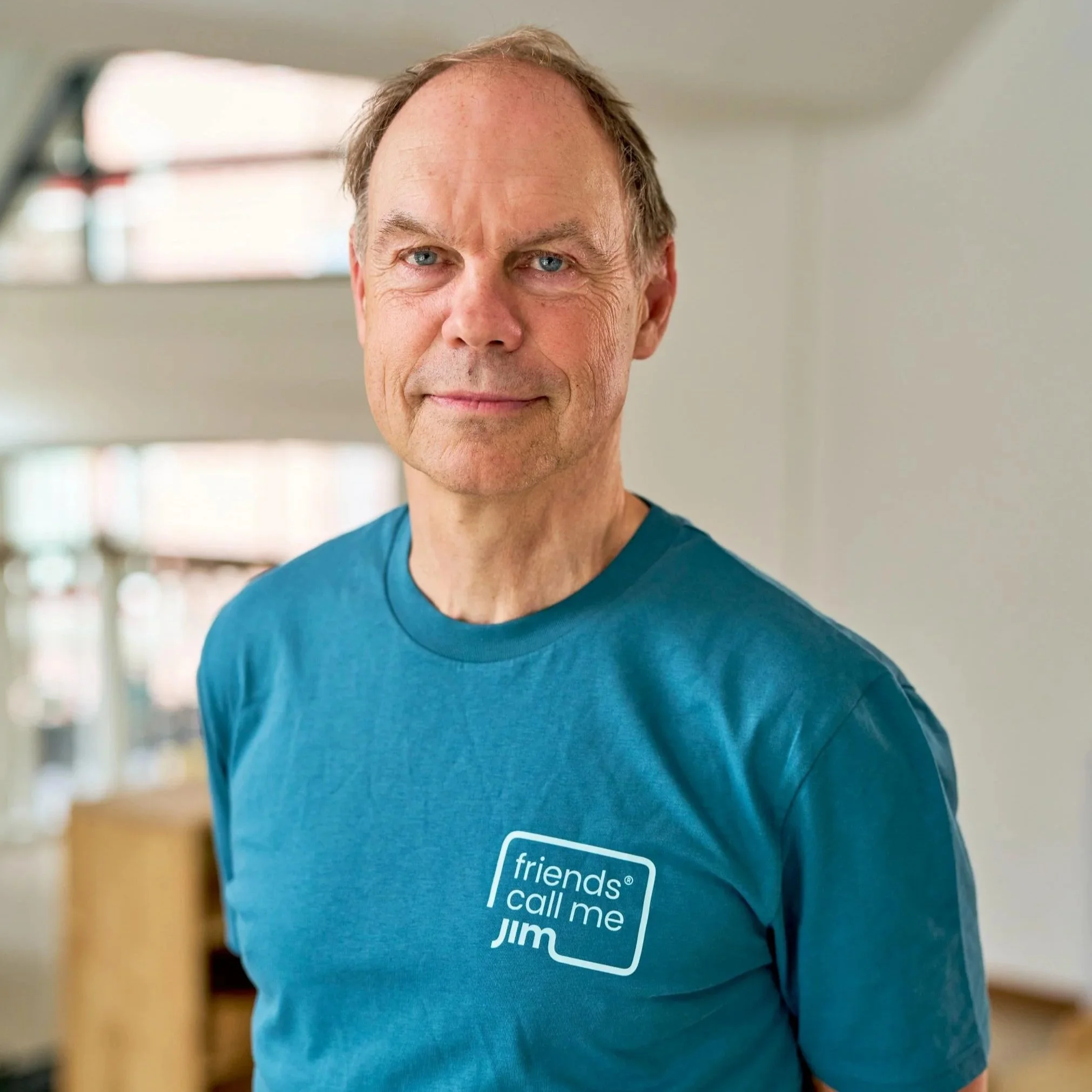 Een oudere man met een blauwe T-shirt met tekst 'friends call me JIM' in een huiselijke binnenruimte.