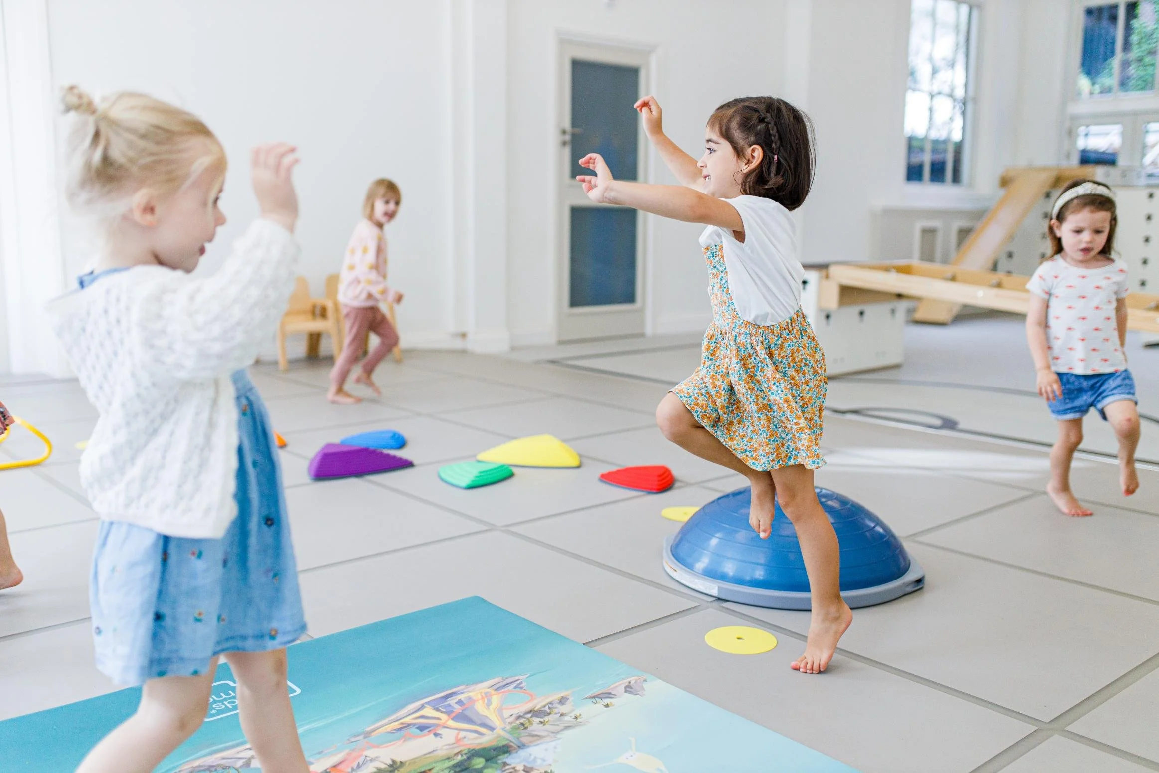 Vijf jonge meisjes spelen en springen in een lichte kleedkamer met gymnastiekmatten en kleurrijk fitnessmateriaal.