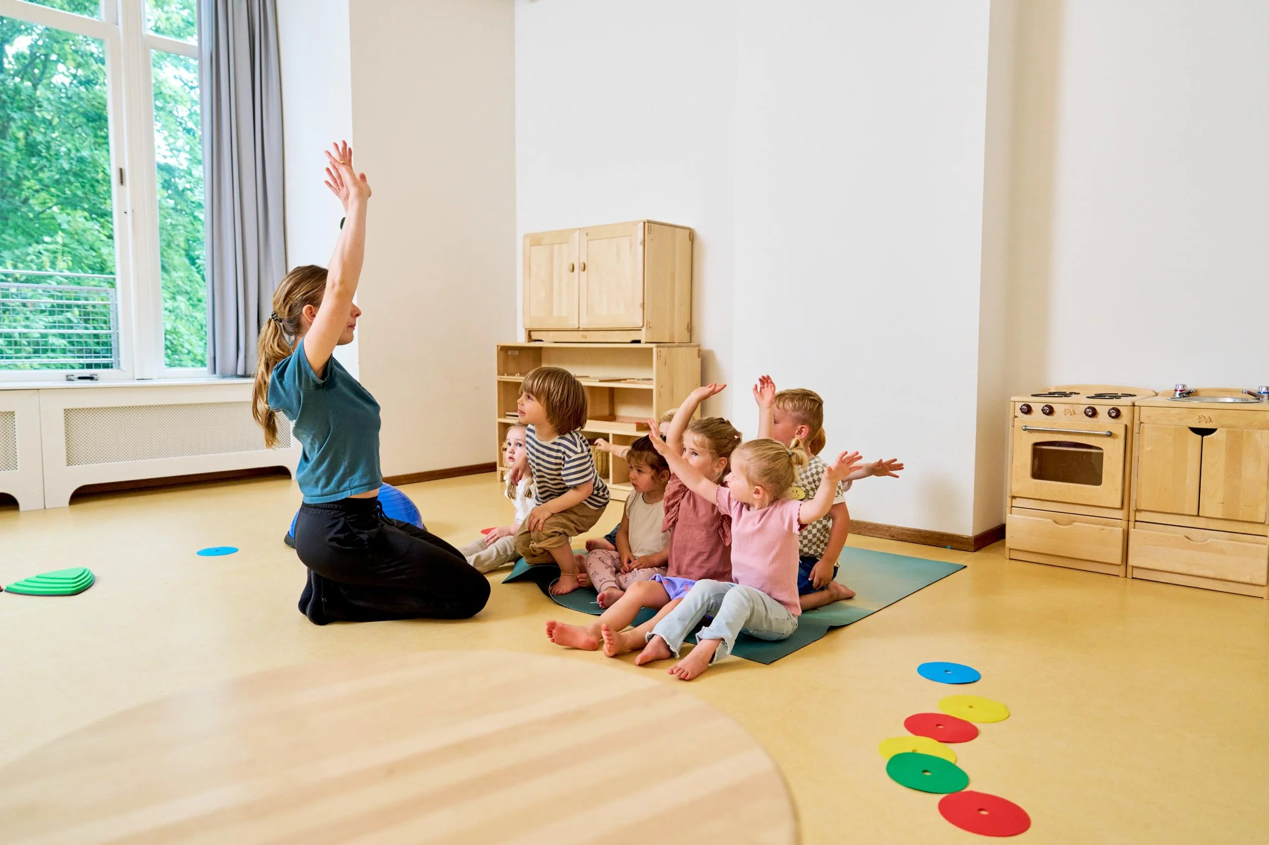 Een jonge vrouw leidt een groepsactiviteit met zeven kleine kinderen in een speelkamer, waarbij ze allemaal hun hand opsteken. De kamer heeft grote ramen en een houten keuken en kast.