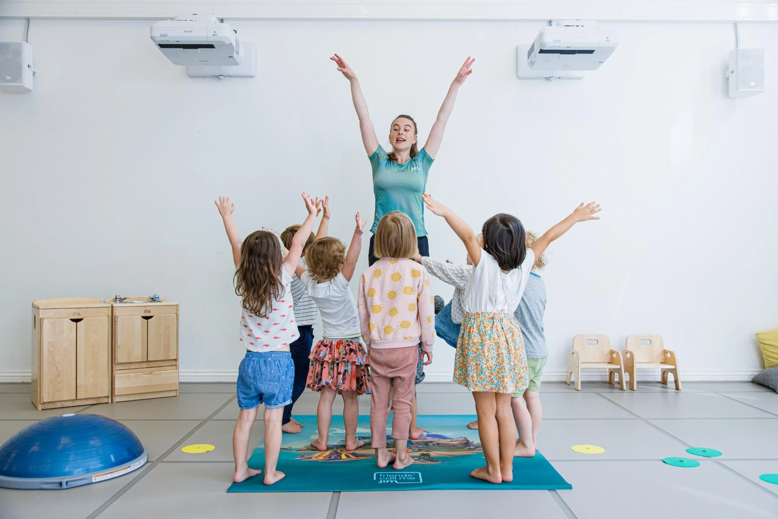Een instructeur geeft een dansles aan jonge kinderen in een gymzaal, iedereen staat op een mat met hun handen omhoog in een vrolijke oefening.
