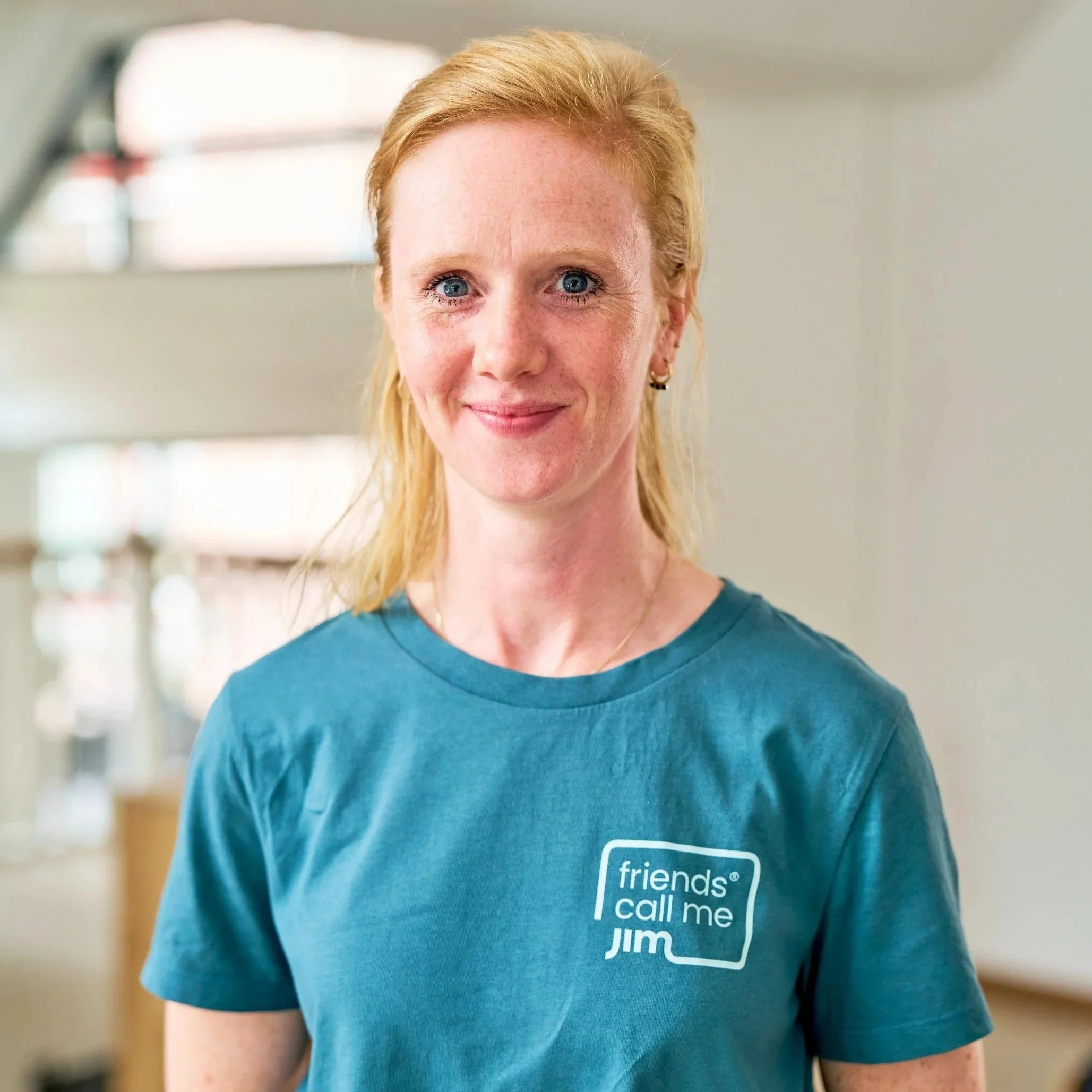 Een vrouw met blond haar en blauwe ogen draagt een turquoise T-shirt met de tekst 'friends call me JIM' op de borst. Ze staat binnen in een lichte, moderne ruimte en glimlacht naar de camera.