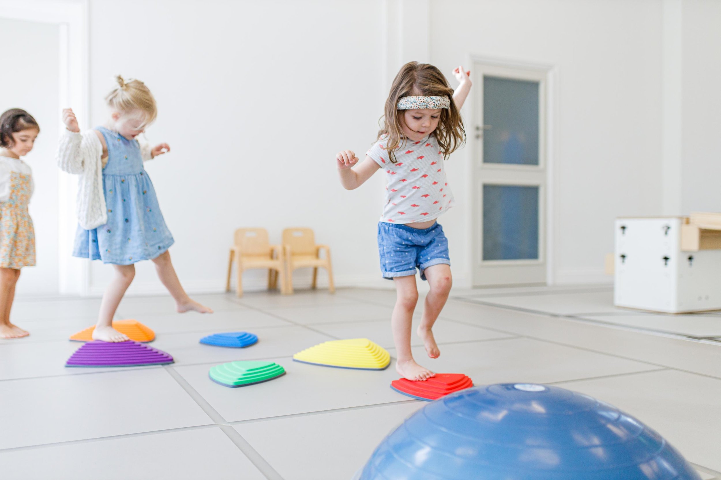 Drie jonge meisjes bewegen op een speelhoogte parcours met gekleurde triangelstenen en een blauwe bal in een witte kamer.
