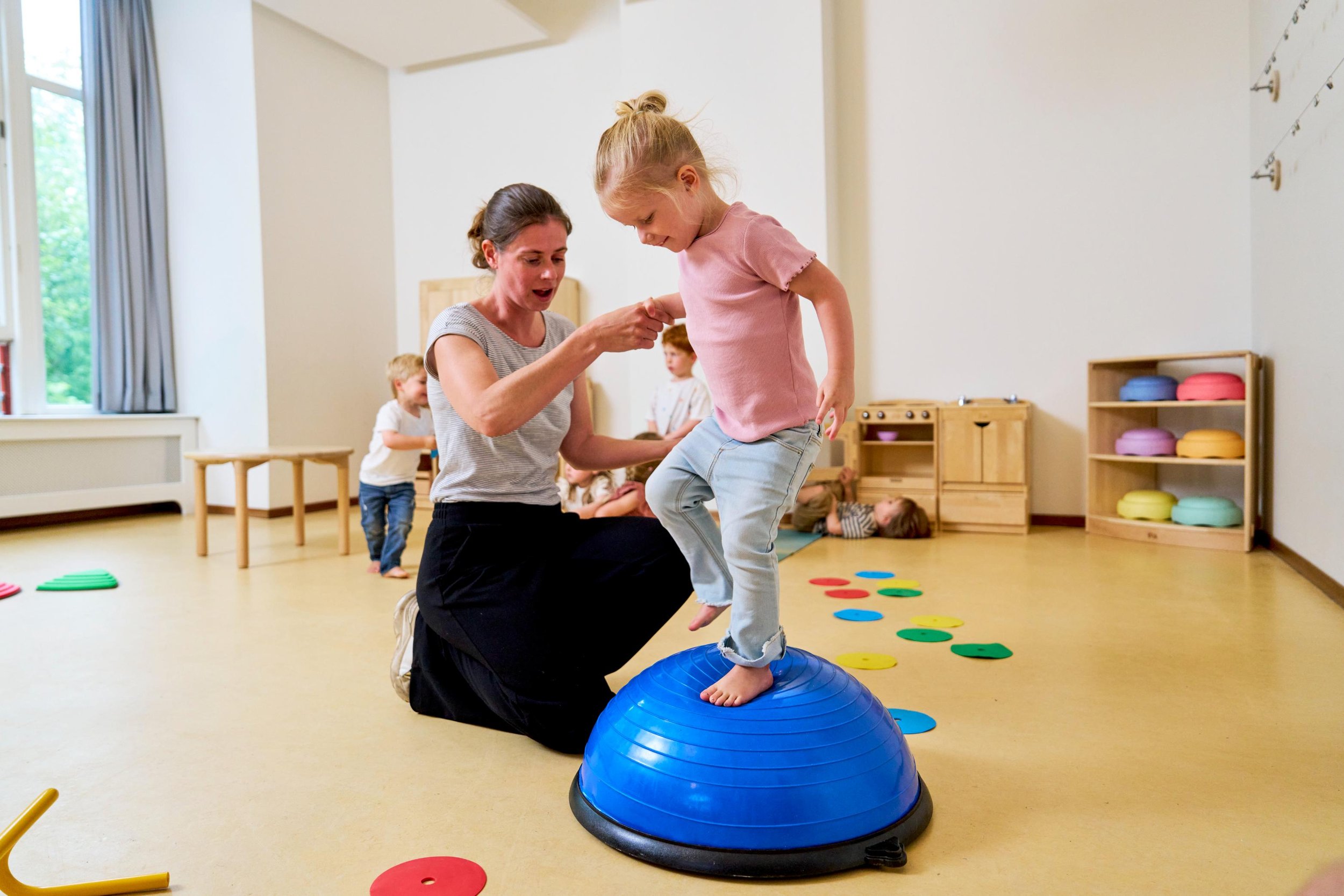 Een jonge meid die op een blauwe evenwichtsbalancebal staat, ondersteund door een vrouw in een speelkamer met andere kinderen op de achtergrond.