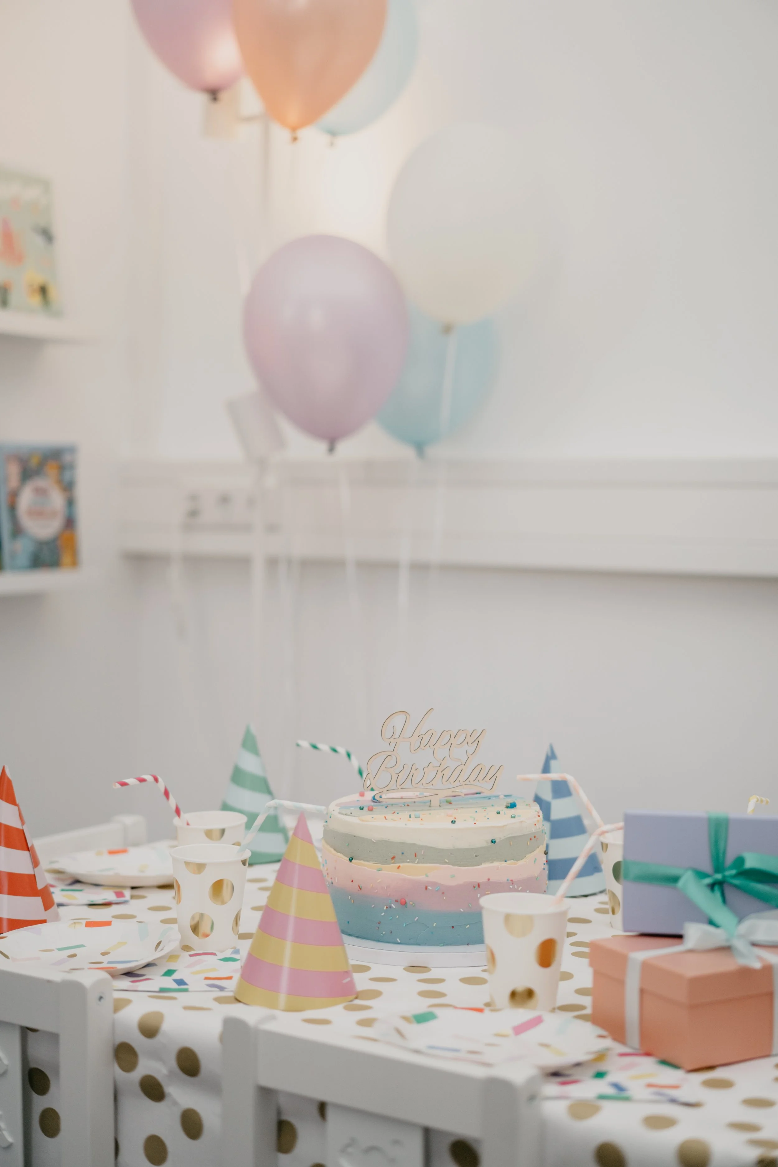 Geburtstagstisch mit bunten Partyhüten, Konfetti, Getränken, einem pastellfarbenen Geburtstagskuchen mit Kerzen, Luftballons im Hintergrund und Geschenken.