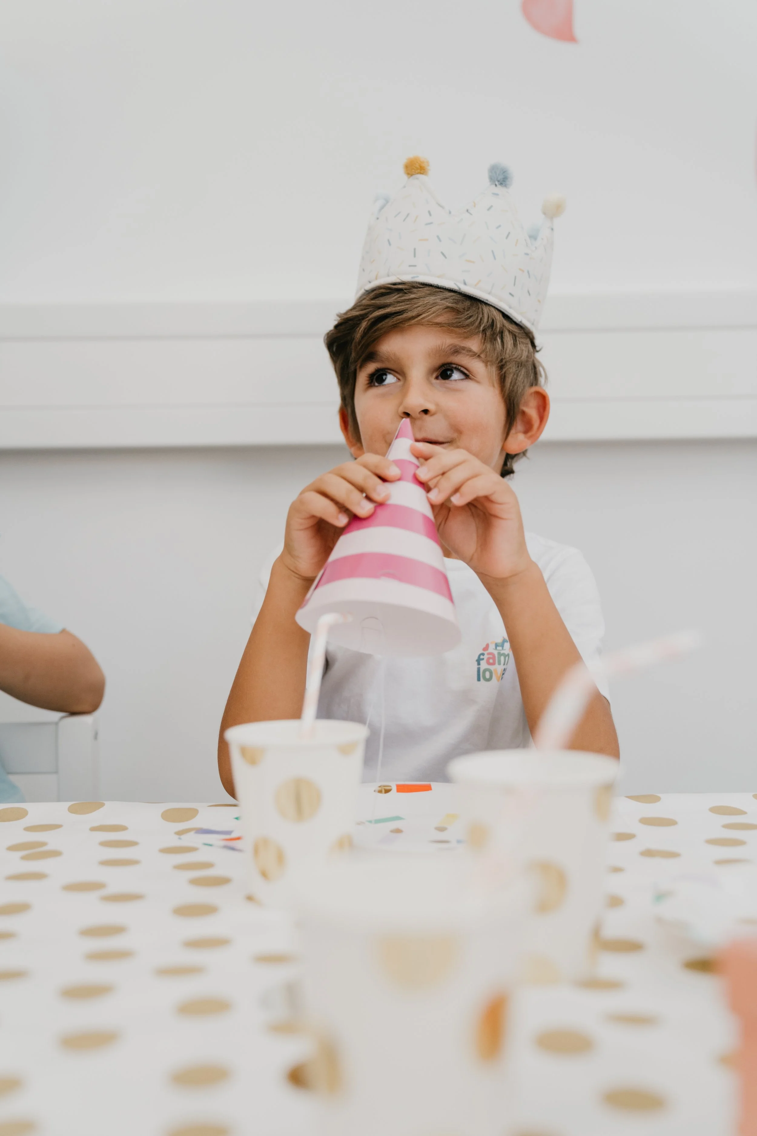 Ein Junge trägt eine Geburtstagskrone und trinkt aus einem Partyhut, es gibt Desserts auf dem Tisch, darunter Becher mit Strohhalmen, bei einer Feier.