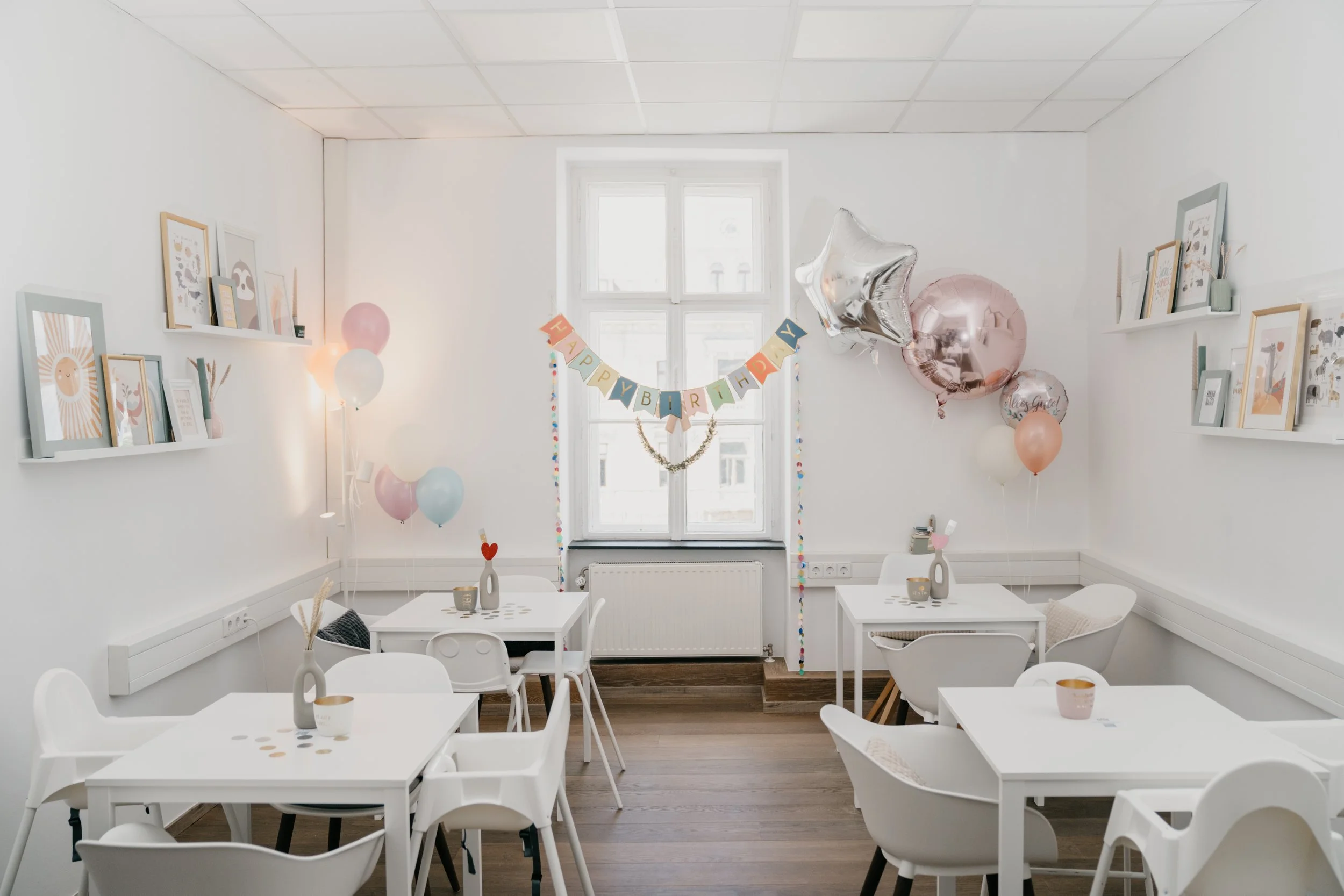 Ein Raum mit weißen Tischen und Stühlen, festliche Dekorationen wie Ballons und eine Geburtstagsbanner, viele Bilder an den Wänden, Fenster und natürliche Beleuchtung.