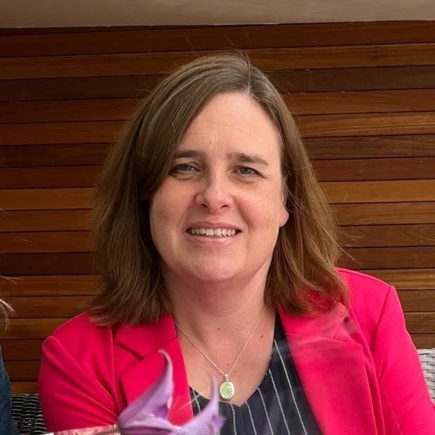 Woman with shoulder-length brown hair wearing a pink blazer and a black and white striped top, sitting outdoors against a wooden panel background.