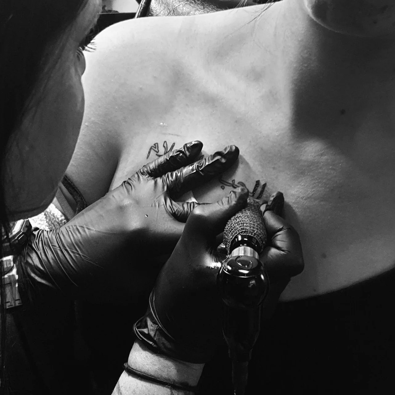 A person with a tattoo on their chest is getting a tattoo done by an artist wearing black gloves, using a tattoo machine.
