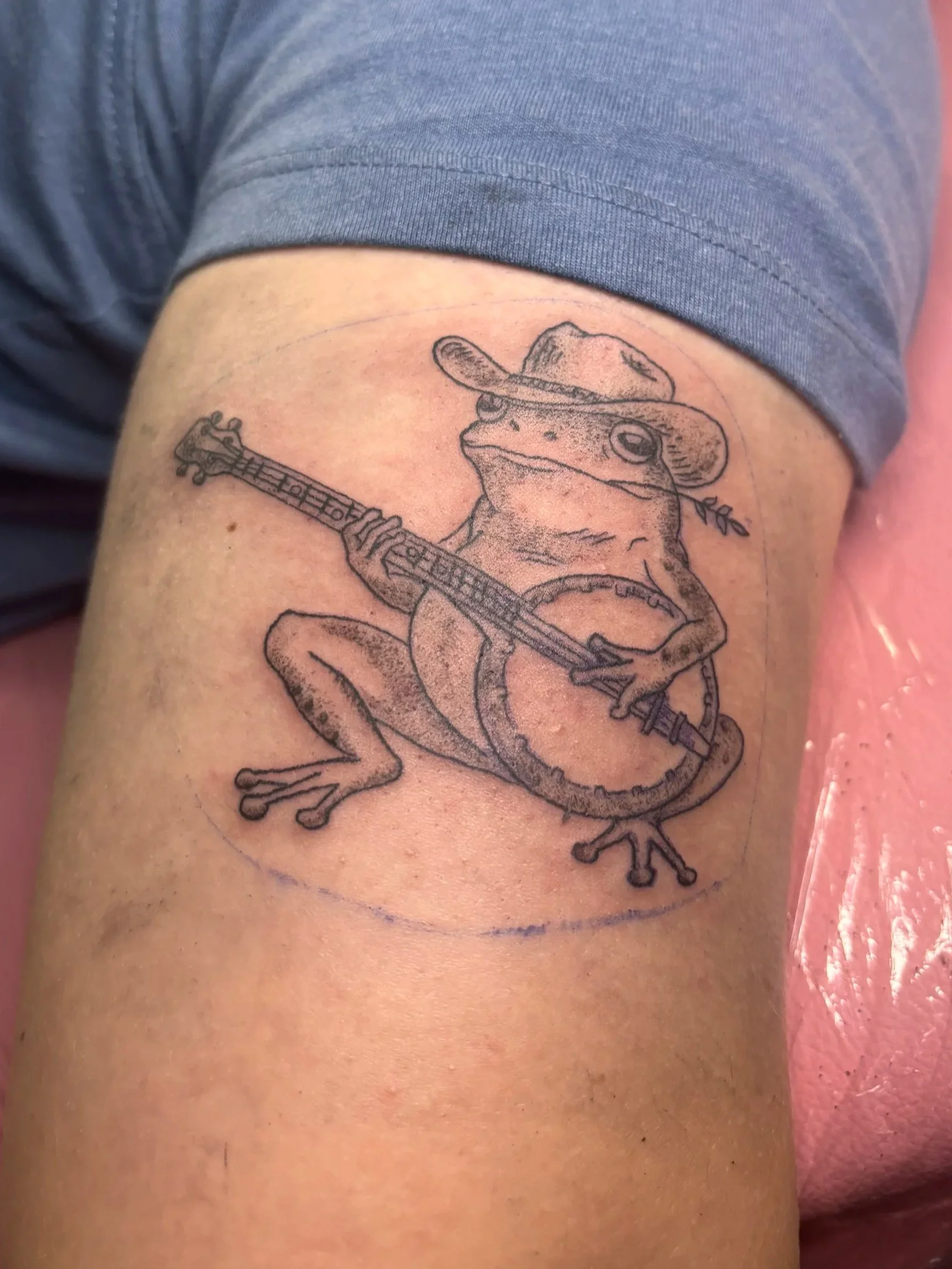 Tattoo of a frog wearing a cowboy hat, playing a guitar, with a lasso coiled around its body.
