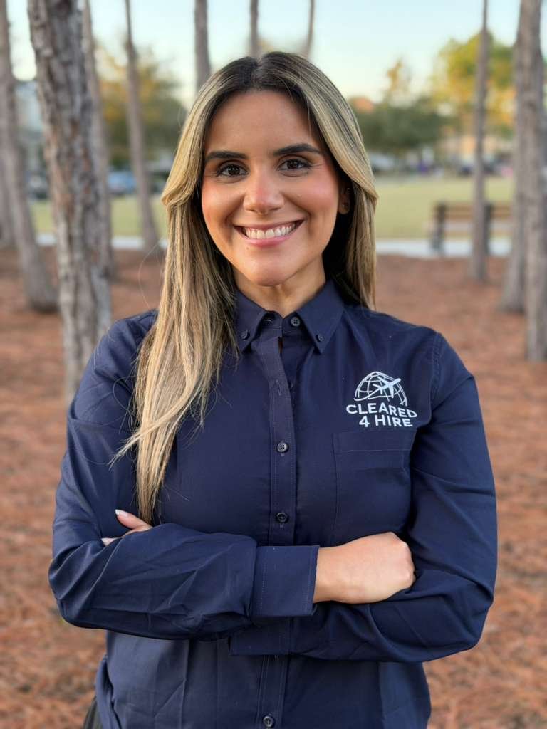 A woman with long blonde hair smiling, standing outside in a wooded park, wearing a navy blue collared shirt with a logo that says 'Cleared 4 Hire' and a globe design.