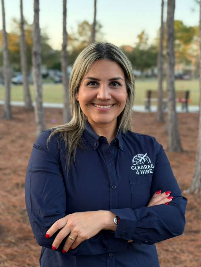 A woman with blonde hair, wearing a navy blue collared shirt with an embroidered logo reading 'Cleared 4 Hire,' standing outdoors among trees, smiling with arms crossed.