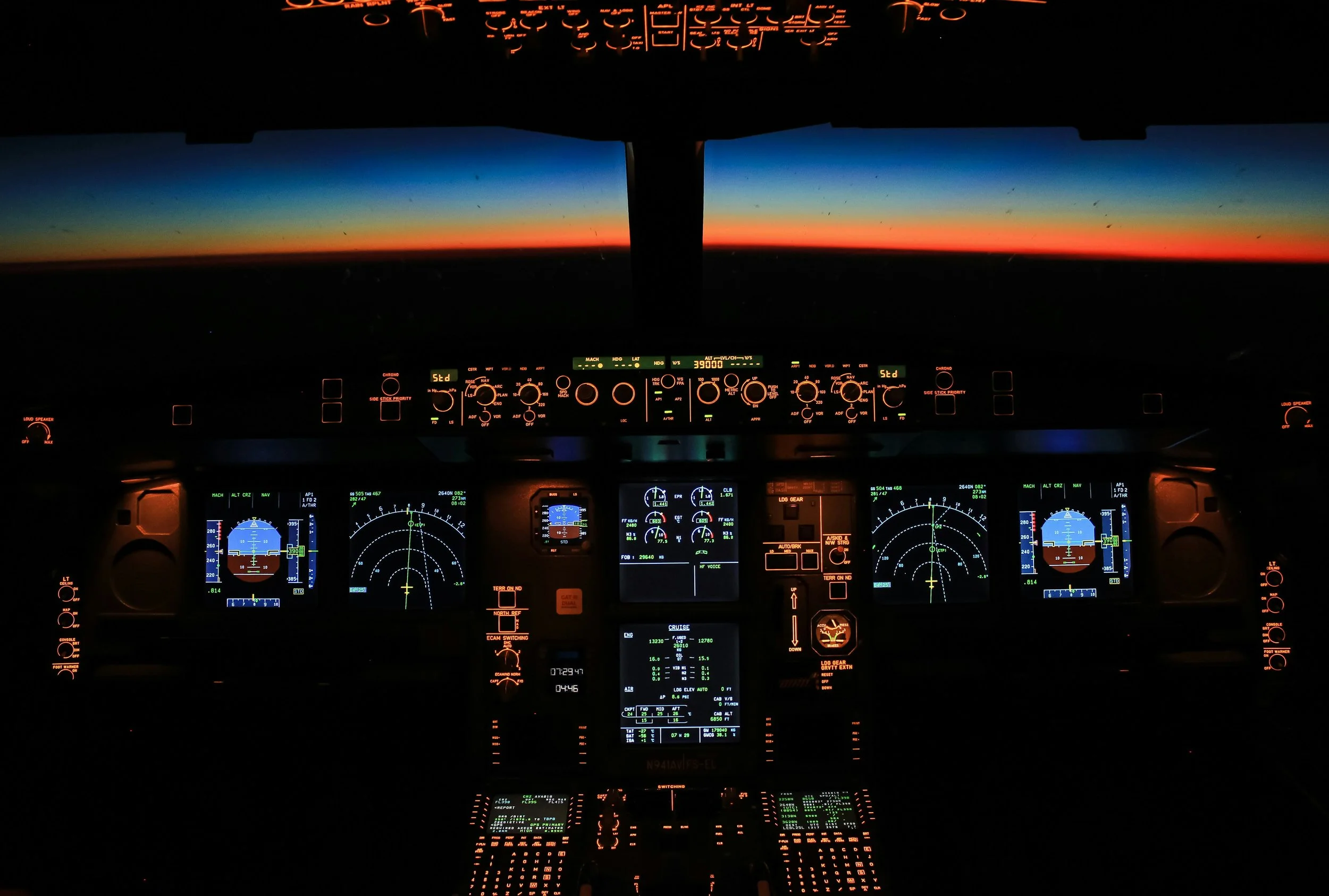 Inside an airplane cockpit during sunset, showing illuminated instrument panels and a view of the colorful sky through the windshield.