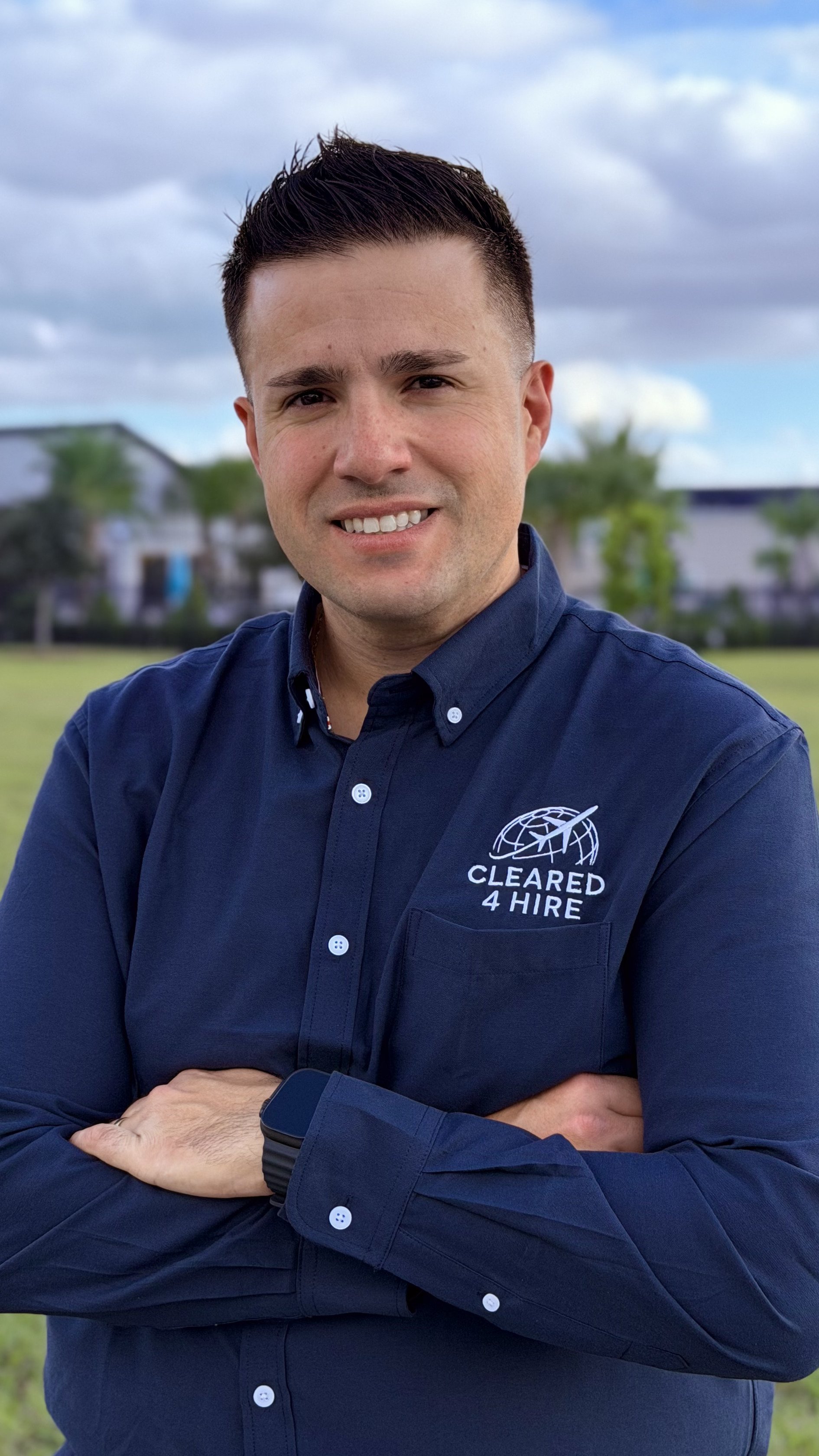 A man with short dark hair wearing a navy blue button-up shirt with a logo and text reading 'Cleared 4 Hire' on it, standing outdoors with trees and buildings in the background.