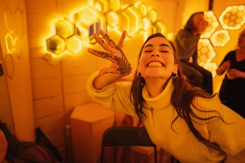 A smiling woman in a yellow sweater making a playful gesture with her hand, holding a decorative insect or jewelry on her fingers, in a warmly lit room with honeycomb-shaped wall decorations.