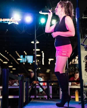 A young woman singing into a microphone on stage with colorful stage lights, wearing a black top, pink shorts, and high boots.