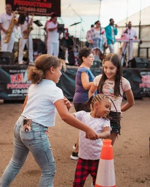 Children playing a game at an outdoor event with a band performing on stage in the background.