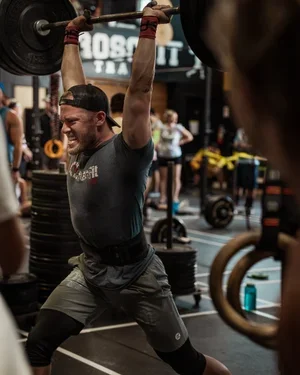 A man lifting a heavy barbell overhead during a CrossFit workout at a gym.