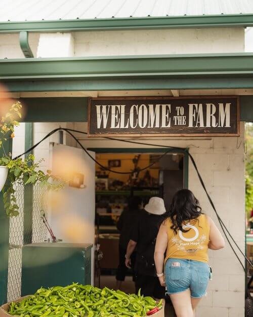 People entering a farm stand or market under a sign that says "Welcome to the Farm," with fresh green peppers displayed at the entrance.