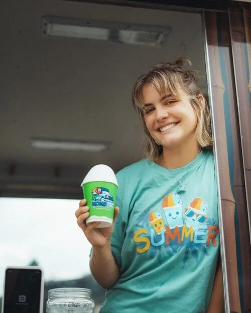 A young woman with blonde hair smiling and holding a small container of ice cream, wearing a blue summer-themed T-shirt, standing near a window inside a building.