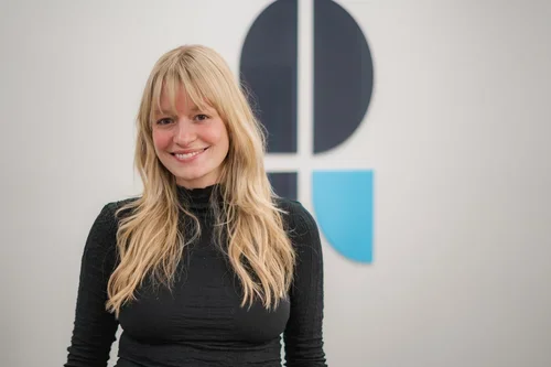 A smiling woman with long blonde hair wearing a black top, standing in front of a wall with a geometric logo.