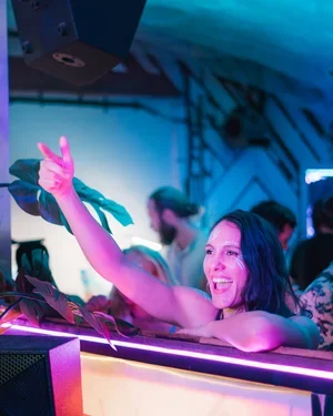 A woman at a party making a peace sign with her hand, smiling, with neon lighting and other people in the background.