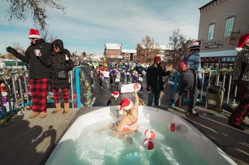 A woman in a Santa hat relaxing in a dog pool with Christmas balls, while people gather outside a store decorated for Christmas.