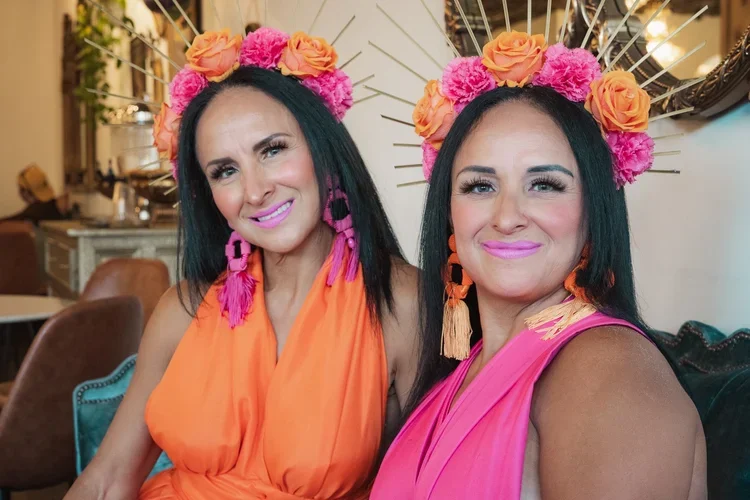 Two women sitting together, wearing colorful floral headbands, bright dresses, and colorful earrings, smiling at the camera in a decorated indoor setting.