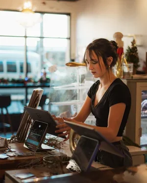 A woman working behind the counter at a cafe or restaurant, using a tablet and a point-of-sale system, with sunlight streaming through large windows in the background.