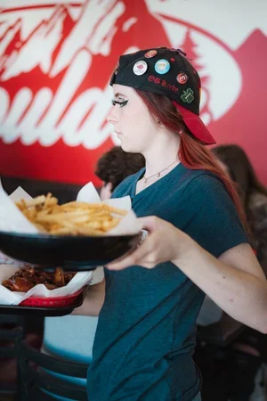 A young woman with red hair wearing a black cap decorated with pins and a blue shirt, holding a tray of French fries at a restaurant.