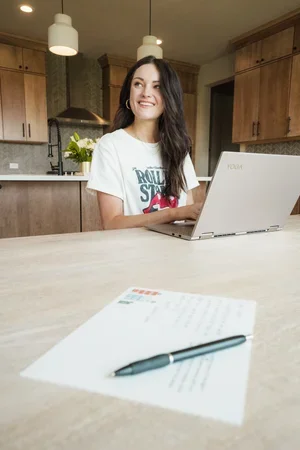 A young woman sitting at a kitchen counter using a laptop, with a notepad and pen in the foreground.