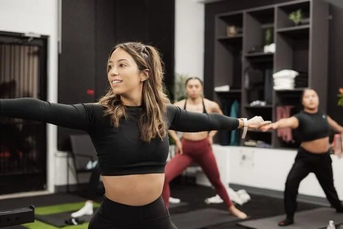 A woman leading a fitness class, with others stretching behind her in a gym.
