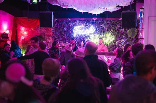 People enjoying live music performance in a dimly lit nightclub with colorful neon lights and decorative greenery.