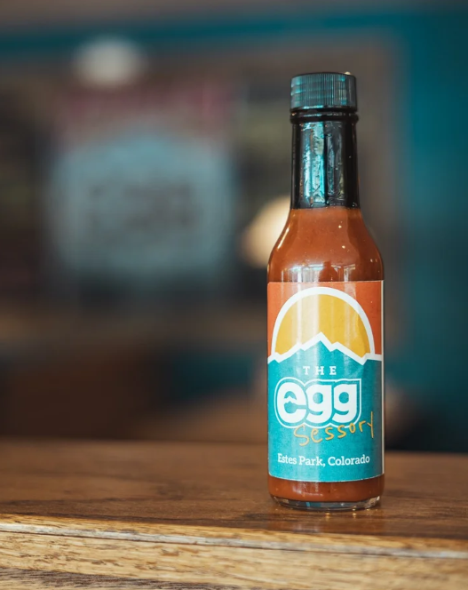 A small hot sauce bottle with a label that reads 'The Egg Sessory' and 'Estes Park, Colorado', placed on a wooden surface with a blurred background.