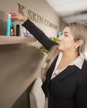 A woman in a black blazer reaching for skincare products on a shelf at a skincare clinic or store.