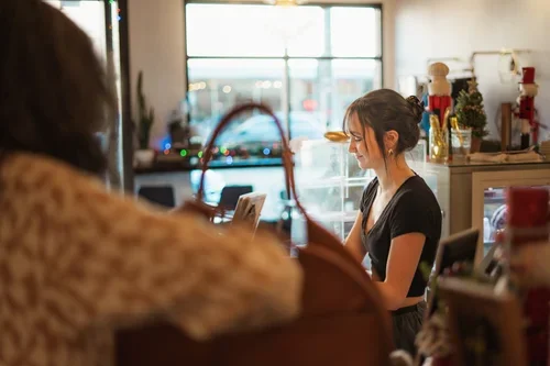 A woman with glasses and a bun hairdo sitting behind a counter in a cozy shop, smiling, with Christmas decorations in the background.