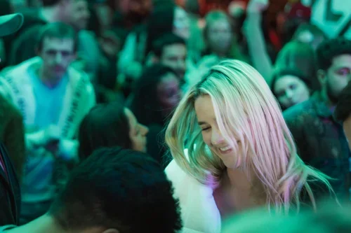 A crowd of diverse people, with a woman with long blonde hair smiling, in a setting with colorful lighting.